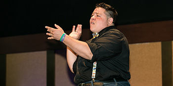 A Latino man uses American Sign Language to communicate. He is wearing a black shirt with suspenders.
