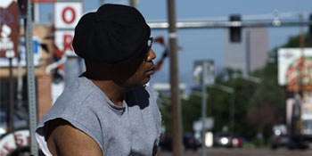 Image of black man in a black beanie hat standing on the sidewalk on a sunny day.