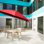 Image of a patio table and two patio chairs with a red patio umbrella inside the enclosed private patio of Sanderson Apartments.