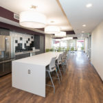 Image of the community room at Sanderson Apartments. There is a white kitchen countertop with gray stools, a refrigerator behind the counter, and a backsplash counter with a sink.
