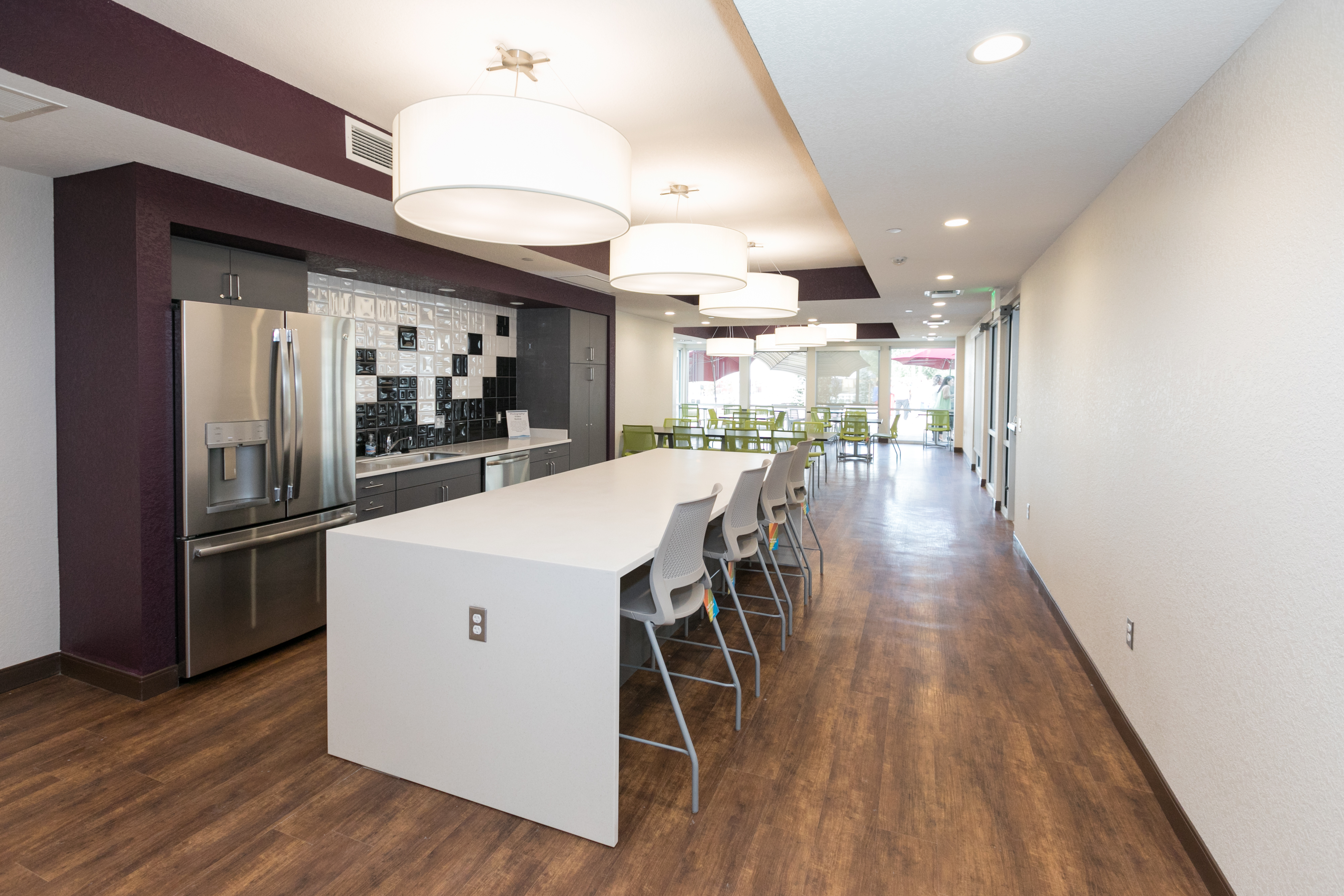 Image of the community room at Sanderson Apartments. There is a white kitchen countertop with gray stools, a refrigerator behind the counter, and a backsplash counter with a sink.