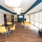 Image of the cafe room at Sanderson Apartments. There are three tables, two of which are square and one is round. The square tables have four yellow chairs around each.