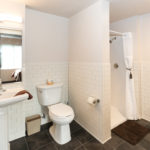 Image of the bathroom in an apartment of Sanderson Apartments. From left to right, there is a pedestal sink with a mirror, a toilet, and a walk-in shower.