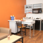 Image of the living room and kitchen in an apartment at Sanderson Apartments. There is a tan couch, counter top with one chair, and kitchen appliances. The wall is orange.