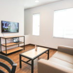 Image of the living room in an apartment in Sanderson Apartments. There is a tan couch, a coffee table, a wall-mounted television, and an entertainment shelf.