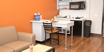 Image of the inside of a one bedroom apartment at Sanderson Apartments. There is a bright orange wall, a white counter with one chair, a tan couch, and a small kitchen in the image.