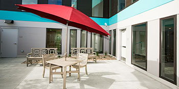 Image of a patio table, two patio chairs, and a red umbrella on the secure patio of Sanderson Apartments.