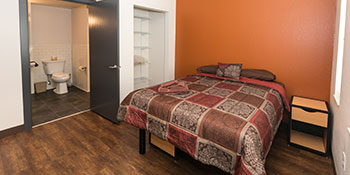 Image of a bed inside a bedroom in Sanderson Apartments. The bedspread is patterned in squares of red, brown, and gold.