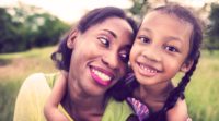 Image of a Black woman and little girl sit together in a meadow. The little girl is to the woman's right and has her arm wrapped around the woman's shoulders.