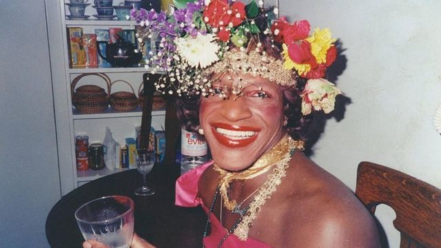 Marsha P. Johnson sits holding a cup. She is wearing a flower crown and smiling widely.