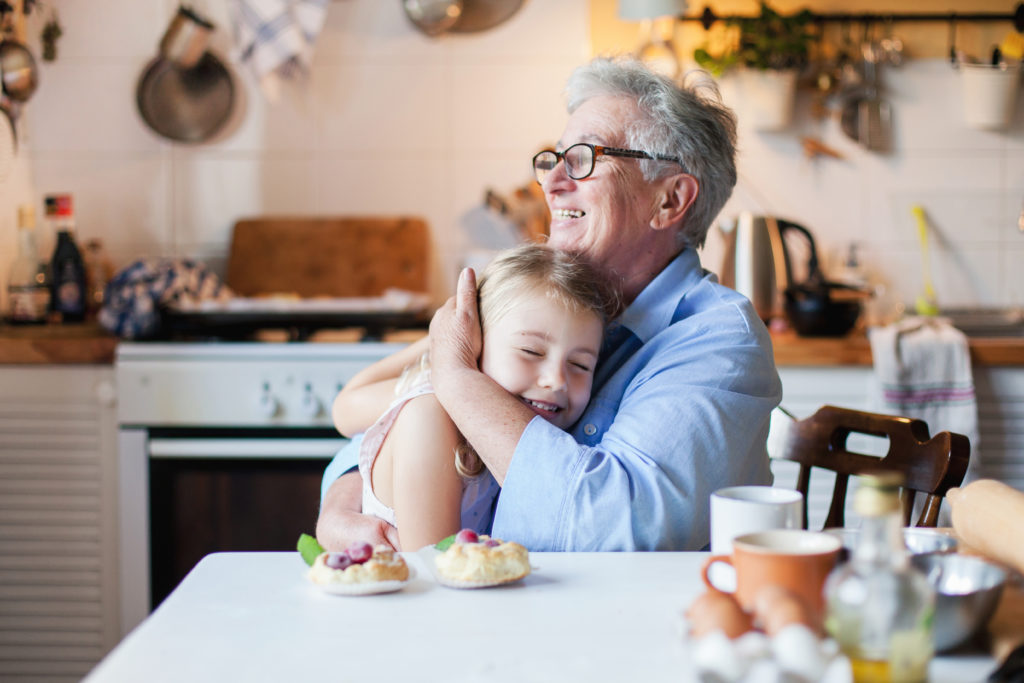 Happy grandmother is hugging granddaughter in cozy home kitchen. Family is cooking together. Senior woman and cute little child girl are smiling. Kid is enjoying kindness, warm hands, care, support.