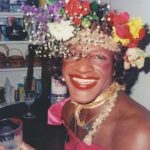 Marsha P. Johnson sits holding a cup. She is wearing a flower crown and smiling widely.