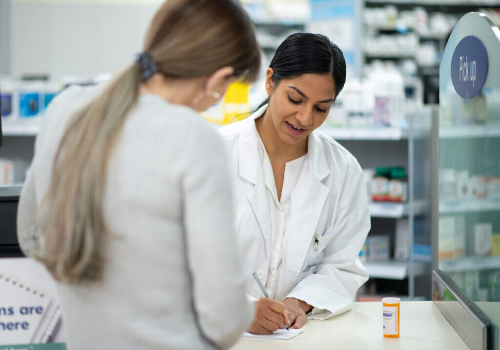 A female pharmacist of Indian decent, stands behind the counter as she reviews a new medication with a customer at the counter.  She is wearing a white lab coat and the medication can be seen sitting on the counter between the two women.