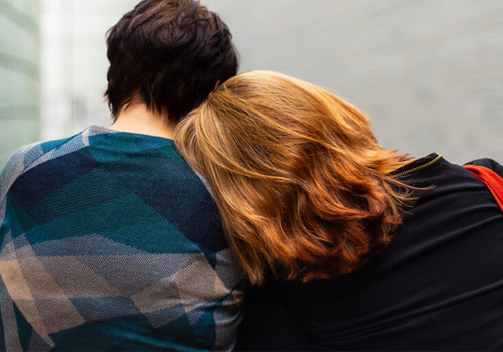 A person with red hair sits to the right of a person with dark brown hair. The image shows them from the back. The person with red hair is leaning their head on the other person's shoulder.