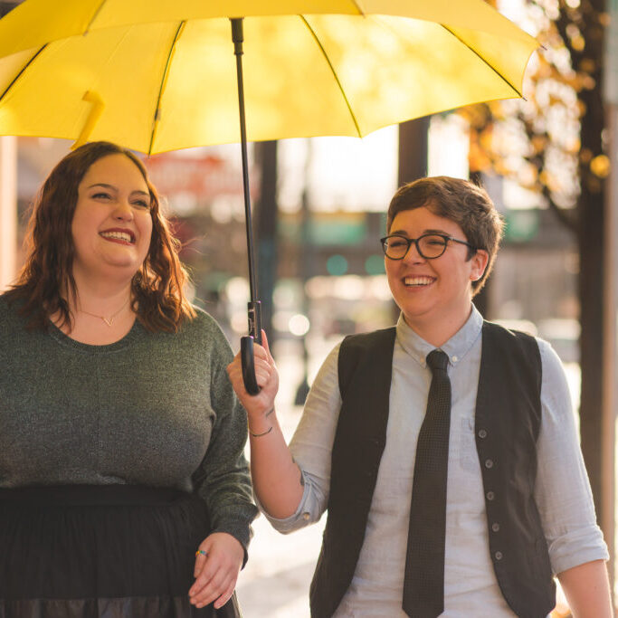 A lesbian couple are  walking out in the city together with an umbrella while laughing