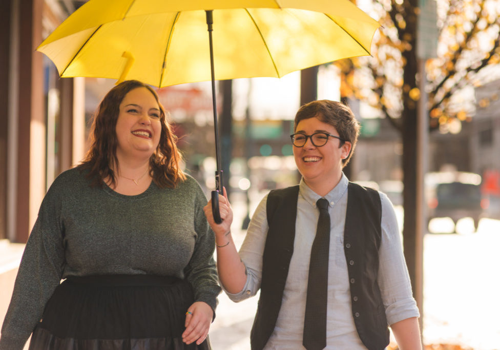 A lesbian couple are  walking out in the city together with an umbrella while laughing