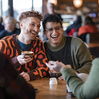 Group of friends sitting in a cafe enjoying a hot drinks together. They are talking, sitting at a table with a view in Tynemouth, North East of England. They are two couples, double dating. One of the couples are LGBTQI+.